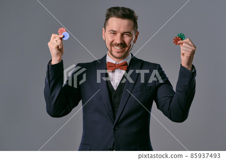 Man in black classic suit and red bow-tie showing some colored chips, posing against gray studio background. Gambling, poker, casino. Close-up. 85937493