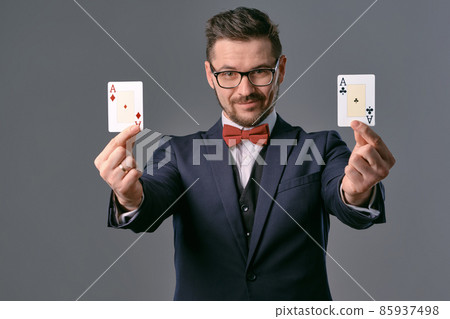 Man in black classic suit, red bow-tie, glases is showing four playing cards, posing on gray studio background. Gambling, poker, casino. Close-up. Man in black classic suit, red bow-tie, glases is showing four playing cards, posing on gray studio background. Gambling, poker, casino. Close-up. 85937498