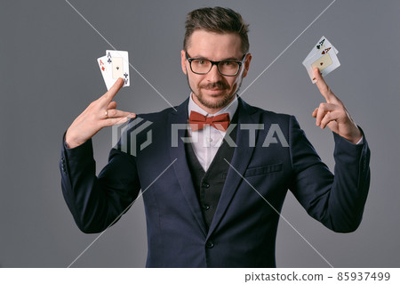 Man in black classic suit, red bow-tie, glases is showing four playing cards, posing on gray studio background. Gambling, poker, casino. Close-up. 85937499