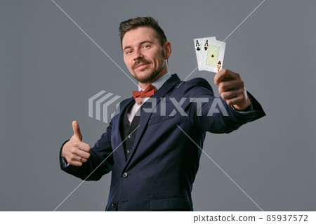 Man in black classic suit and red bow-tie showing two playing cards while posing against gray studio background. Gambling, poker, casino. Close-up. 85937572