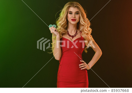 Blonde female in red fitting dress and necklace. She smiling, showing two green chips, posing on colorful background. Poker, casino. Close-up Blonde female in red fitting dress and necklace. She smiling, showing two green chips, posing on colorful background. Poker, casino. Close-up 85937668