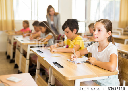 Concentrated tween girl listening to schoolteacher and writing in notebook Concentrated tween girl listening to schoolteacher and writing in notebook 85938116
