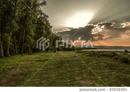 The evening sky with a red sunset from huge clouds, over a forest belt with a clearing next to the shore of Lake Sevan in Armenia. 85939303