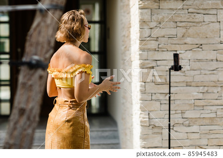 A beautiful girl records her daily blog video on a smartphone mounted on a tripod. Side view. A beautiful girl records her daily blog video on a smartphone mounted on a tripod. Side view. 85945483