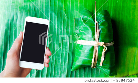 Blank touchscreen holding in hand with bundle of bananas with Sticky Rice on banana leaves concept for using smart device to find how to make dessert 85947524
