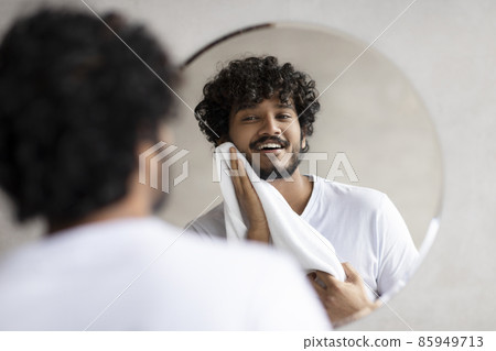 Everyday routine concept. Excited indian man wiping his face with towel while standing in bathroom, looking in mirror Everyday routine concept. Excited indian man wiping his face with towel while standing in bathroom, looking in mirror 85949713