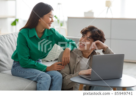 Positive millennial Asian couple using laptop computer, smiling at each other, browsing internet together at home Positive millennial Asian couple using laptop computer, smiling at each other, browsing internet together at home 85949741