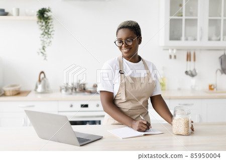 Cheerful millennial pretty black female in glasses, apron writes recipe looks pc in minimalist kitchen interior 85950004