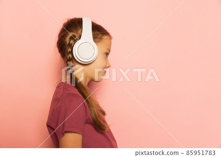 Profile view of little girl wearing t-shirt listening to music in headphones isolated on pink background. Concept of childhood, emotions, study 85951783