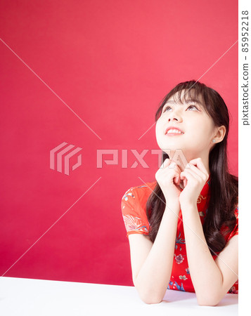 Beautiful asian woman wearing traditional cheongsam qipao dress with hands to her chin, looking to the side on red background. Beautiful asian woman wearing traditional cheongsam qipao dress with hands to her chin, looking to the side on red background. 85952218