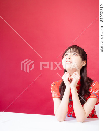 Beautiful asian woman wearing traditional cheongsam qipao dress with hands to her chin, looking to the side on red background. 85952219