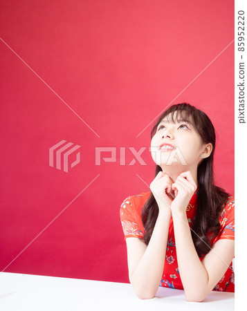 Beautiful asian woman wearing traditional cheongsam qipao dress with hands to her chin, looking to the side on red background. Beautiful asian woman wearing traditional cheongsam qipao dress with hands to her chin, looking to the side on red background. 85952220
