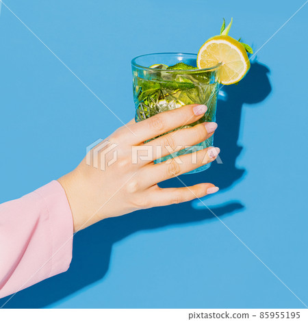 Female hand holding glass with cocktail mojito with lemon isolated on light blue neon background. Concept of taste, alcoholic drinks Female hand holding glass with cocktail mojito with lemon isolated on light blue neon background. Concept of taste, alcoholic drinks 85955195