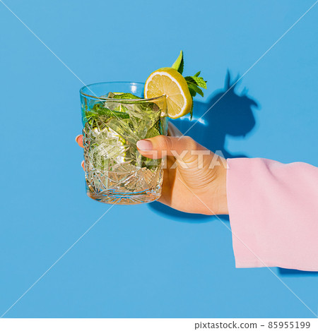 Female hand holding glass with cocktail mojito with lemon isolated on light blue neon background. Concept of taste, alcoholic drinks 85955199