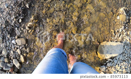 Girl walking barefoot on stones of shallow river close-up, sunny summer day Girl walking barefoot on stones of shallow river close-up, sunny summer day 85973113