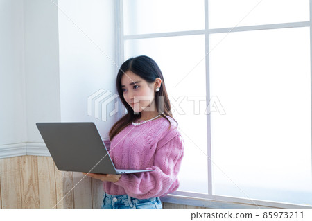 一個年輕的女人站在窗邊看著電腦 一個年輕的女人站在窗邊看著電腦 85973211