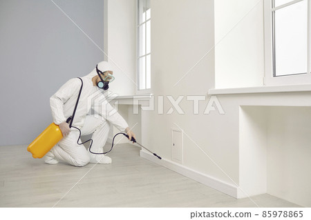 Pest control service guy working inside the house. Man in white protective overalls crouching near wall and spraying cockroach insecticide from yellow sprayer for safe living environment at home 85978865