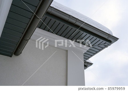 Corner of the house with new gray metal tile roof and rain gutter covered by snow at winter. Metallic Guttering System, Guttering and Drainage Pipe Exterior 85979959