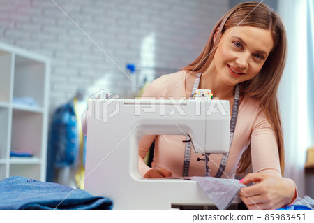Cheerful young seamstress sitting at working table in her office Cheerful young seamstress sitting at working table in her office 85983451