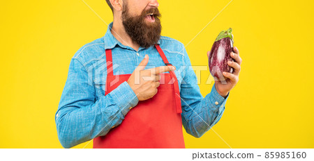 Happy man crop view red apron pointing finger at eggplant yellow background, shopkeeper 85985160