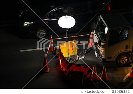 Night construction of the main road A floodlight that illuminates the manhole and a car that passes by Night construction of the main road A floodlight that illuminates the manhole and a car that passes by 85986321