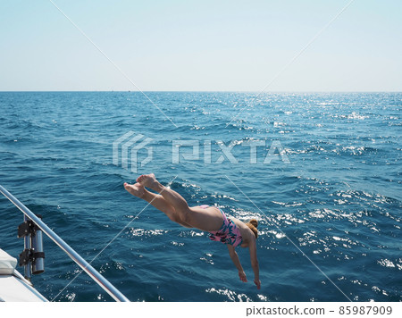A girl dives from a yacht into the open blue sea illuminated by sun glare on a clear day 85987909