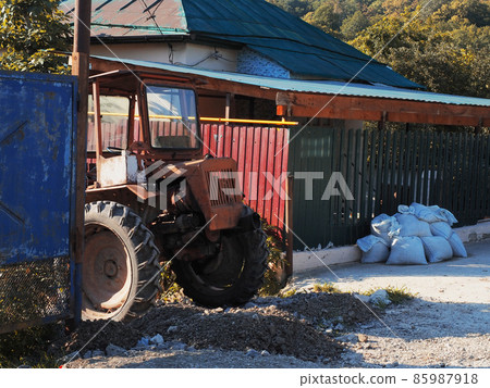 An old red tractor stands on a broken rocky road near an old fenced private house 85987918