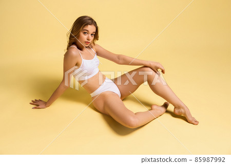 Top view of young slim girl, blonde in underwear sitting on floor isolated over yellow studio background. Natural beauty concept. Top view of young slim girl, blonde in underwear sitting on floor isolated over yellow studio background. Natural beauty concept. 85987992
