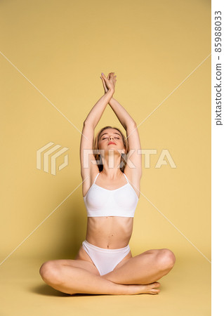Studio shot of young adorable woman in underwear sitting on floor isolated over yellow background. Natural beauty concept. Studio shot of young adorable woman in underwear sitting on floor isolated over yellow background. Natural beauty concept. 85988033