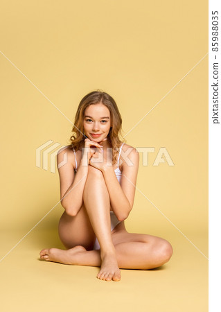 Studio shot of young adorable woman in underwear sitting on floor isolated over yellow background. Natural beauty concept. Studio shot of young adorable woman in underwear sitting on floor isolated over yellow background. Natural beauty concept. 85988035