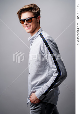 Portrait of smiling handsome guy standing in sunglasses holds arms in pockets, isolated on a gray background Portrait of smiling handsome guy standing in sunglasses holds arms in pockets, isolated on a gray background 85990859