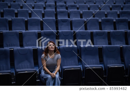 Caucasian red-haired woman sits on the front row in a cinema in an empty hall. The girl is watching a movie alone.  85993482