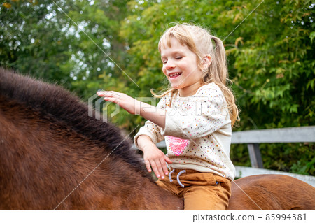 cheerful girl sits astride and strokes a horse on the mane 85994381