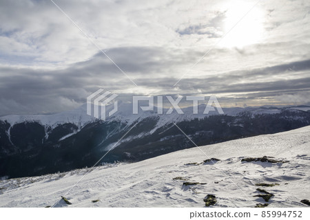 Winter mountain landscape. Wide view of distant woody mountain peaks covered with shiny snow, ice and glaciers under cloudy blue sky on clear cold sunny winter day. 85994752