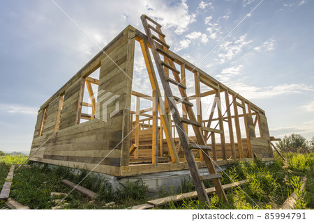 Unfinished ecological house under construction in green field on blue sky background. Trenches filled with cement and wooden walls with window openings of future comfortable cottage in quiet area. Unfinished ecological house under construction in green field on blue sky background. Trenches filled with cement and wooden walls with window openings of future comfortable cottage in quiet area. 85994791