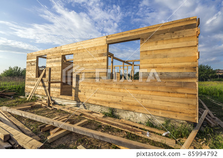 Unfinished ecological house under construction in green field on blue sky background. Trenches filled with cement and wooden walls with window openings of future comfortable cottage in quiet area. Unfinished ecological house under construction in green field on blue sky background. Trenches filled with cement and wooden walls with window openings of future comfortable cottage in quiet area. 85994792