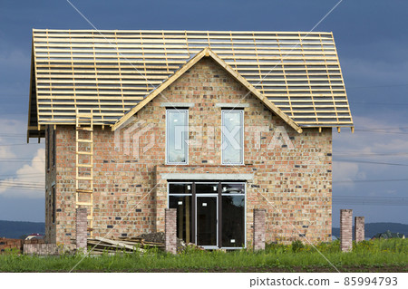 Two-story residential house with plastic windows, wide door and wooden frame for roof under construction. High wooden ladder at brick walls and brick columns for fence on dark blue sky background. Two-story residential house with plastic windows, wide door and wooden frame for roof under construction. High wooden ladder at brick walls and brick columns for fence on dark blue sky background. 85994793