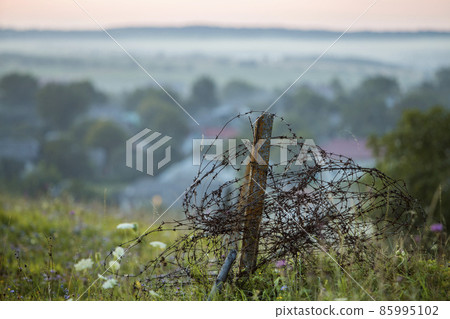 Old big barbed wire coiled on rusty pole, broken garden fence on grassy blooming hill on misty light sky at dawn or evening and blue foggy blurred houses and gardens deep below copy space background. 85995102