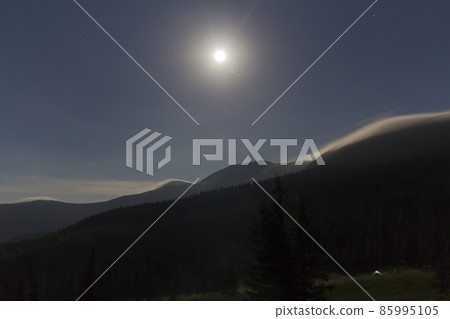 Night view of bright moon shining in dark blue sky over mountains covered with evergreen forest and small tourist tent on grassy valley. Beauty and magic of nature, tourism and traveling concept. Night view of bright moon shining in dark blue sky over mountains covered with evergreen forest and small tourist tent on grassy valley. Beauty and magic of nature, tourism and traveling concept. 85995105