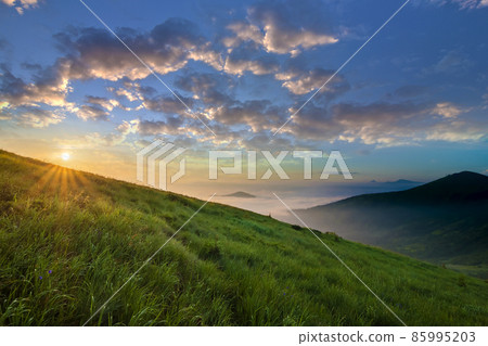 Mountain landscape in nice weather at sunrise. Green grassy steep hill, foggy valley and distant mountains under bright blue sky with lit by raising sun white clouds. Beauty of nature concept. 85995203