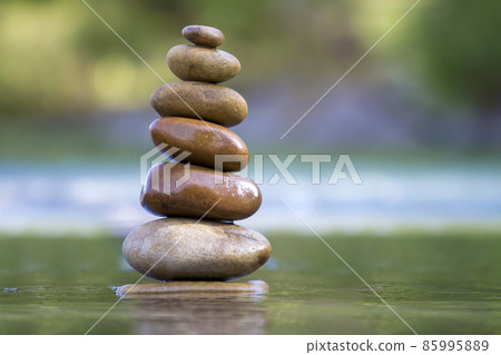 Close-up abstract image of wet rough natural brown uneven different sizes and forms stones balanced like pyramid pile landmark in shallow water on blurred blue-green misty copy space background. 85995889