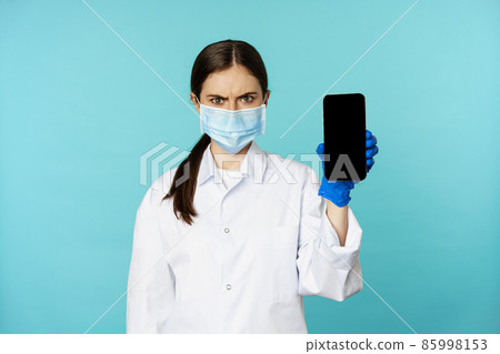 Angry and confused woman doctor in face mask, gloves, showing smartphone app, mobile screen, frowning upset, standing over blue background 85998153