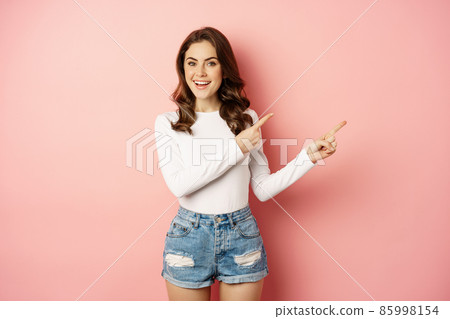 Happy smiling beautiful woman, pointing fingers right, demonstrating banner, company logo on empty space, promo text, standing over pink background 85998154