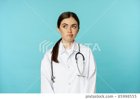 Image of woman doctor, female medial staff in white lab coat, looking away thoughful, making decision, thinking of smth, standing over blue background Image of woman doctor, female medial staff in white lab coat, looking away thoughful, making decision, thinking of smth, standing over blue background 85998555