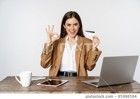 Portrait of female entrepreneur sitting at office table, showing credit card and thumbs up, recommending, standing over white background Portrait of female entrepreneur sitting at office table, showing credit card and thumbs up, recommending, standing over white background 85998584