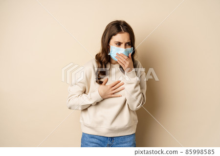 Covid-19 and health concept. Sick woman in medical face mask coughing, feeling ill with sour throat, standing over beige background Covid-19 and health concept. Sick woman in medical face mask coughing, feeling ill with sour throat, standing over beige background 85998585