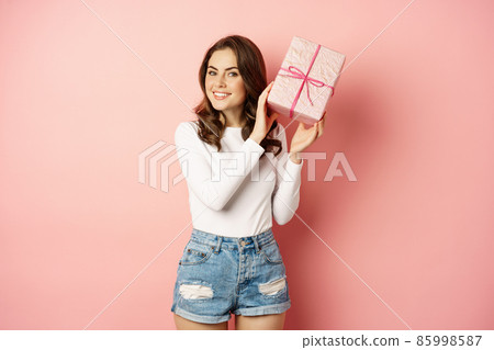 Cute smiling girl guessing whats inside pink present box, shaking gift and laughing, standing against studio background Cute smiling girl guessing whats inside pink present box, shaking gift and laughing, standing against studio background 85998587