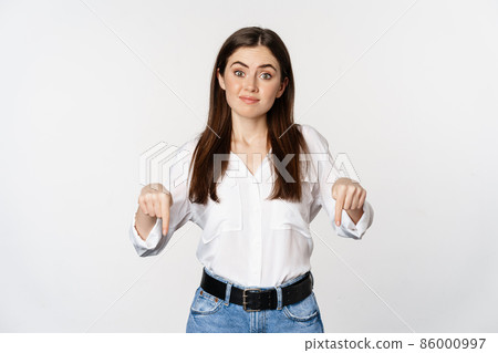 Doubtful cute girl shrugging unsure, smiling and pointing fingers down, standing over white background Doubtful cute girl shrugging unsure, smiling and pointing fingers down, standing over white background 86000997