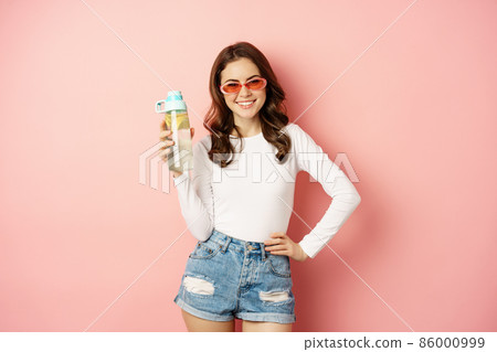 Stylish girl in spring outfit, wearing sunglasses, holding water bottle with lemon, healthy drink, laughing and smiling, standing over pink background Stylish girl in spring outfit, wearing sunglasses, holding water bottle with lemon, healthy drink, laughing and smiling, standing over pink background 86000999