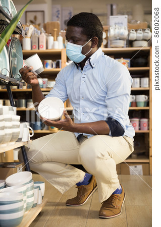 Man in protective mask carefully chooses ceramic mugs in tableware store Man in protective mask carefully chooses ceramic mugs in tableware store 86002068